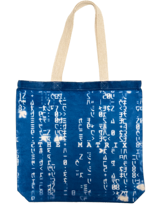 A blue fabric tote bag with white Matrix characters printed vertically across the front, featuring two beige canvas shoulder straps.